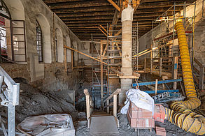 Eine Großbaustelle im Innenraum einer großen historischen Halle ist zu sehen. In der Mitte befindet sich eine große Steinsäule um die ein Gerüst aufgebaut ist. Der Keller wurde ausgegraben und man sieht die Tiefe. Im rechten Bildrand befindet sich ein langer, gelber Bauschutt-Schlauch, der von einem Gerüst herunterführt.