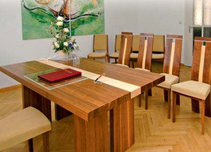 Im Vordergrund steht ein dunkler Holztisch mit einem beigen Streifen auf der Tischplatte. Ein Blumengesteck und eine rote Mappe sowie ein Füller befinden sich auf dem Tisch. Dahinter sieht man Stuhlreihen für das Brautpaar und die Gäste. An der Wand hängt ein großformatiges buntes Gemälde.